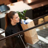A person riding an escalator while carrying a large brown paper shopping bag filled with items, including a green potted plant and folded clothing.