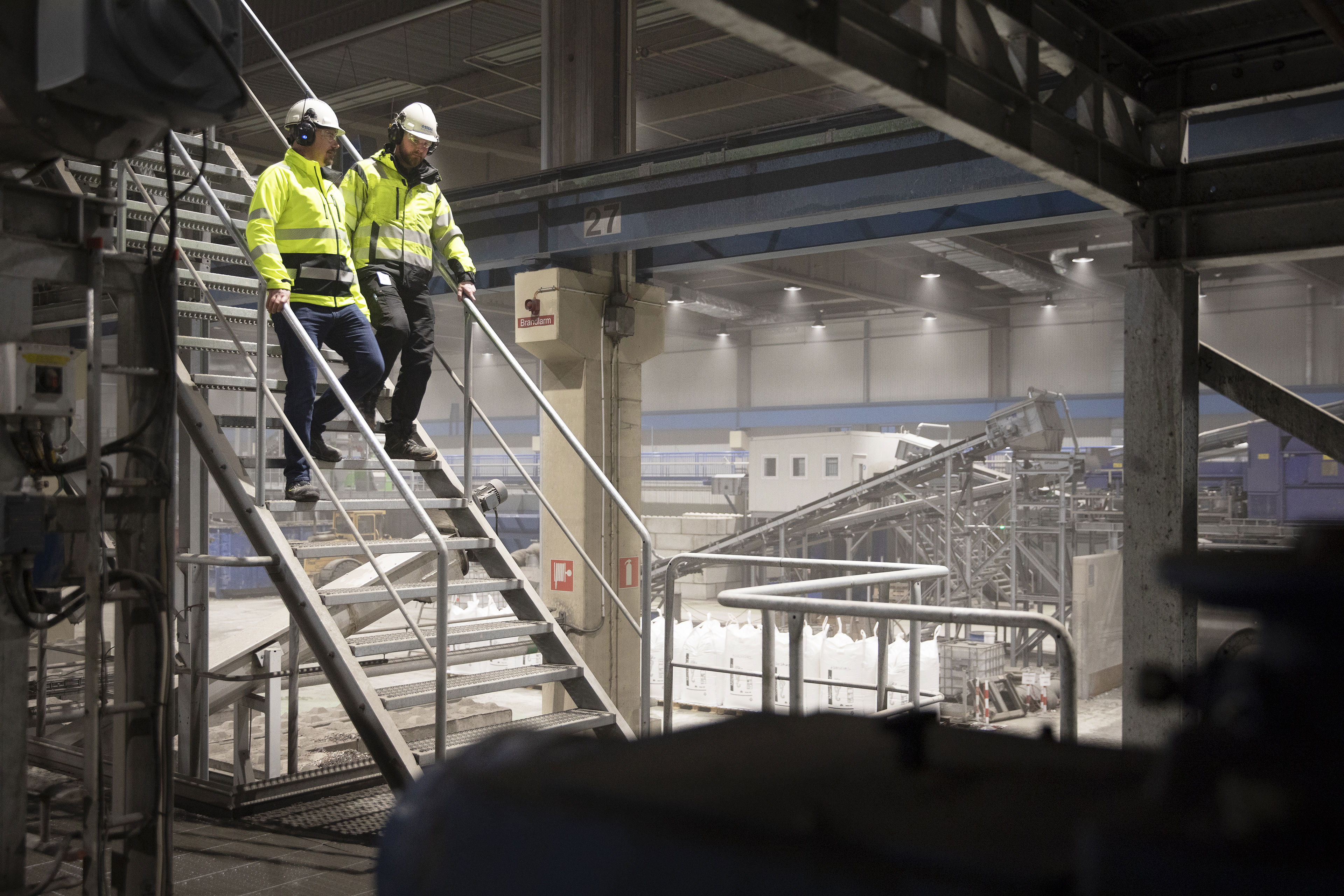 Two male Stena Recycling employees in hi-visibility jackets and protective helmets walk down stairs in an industrial facility