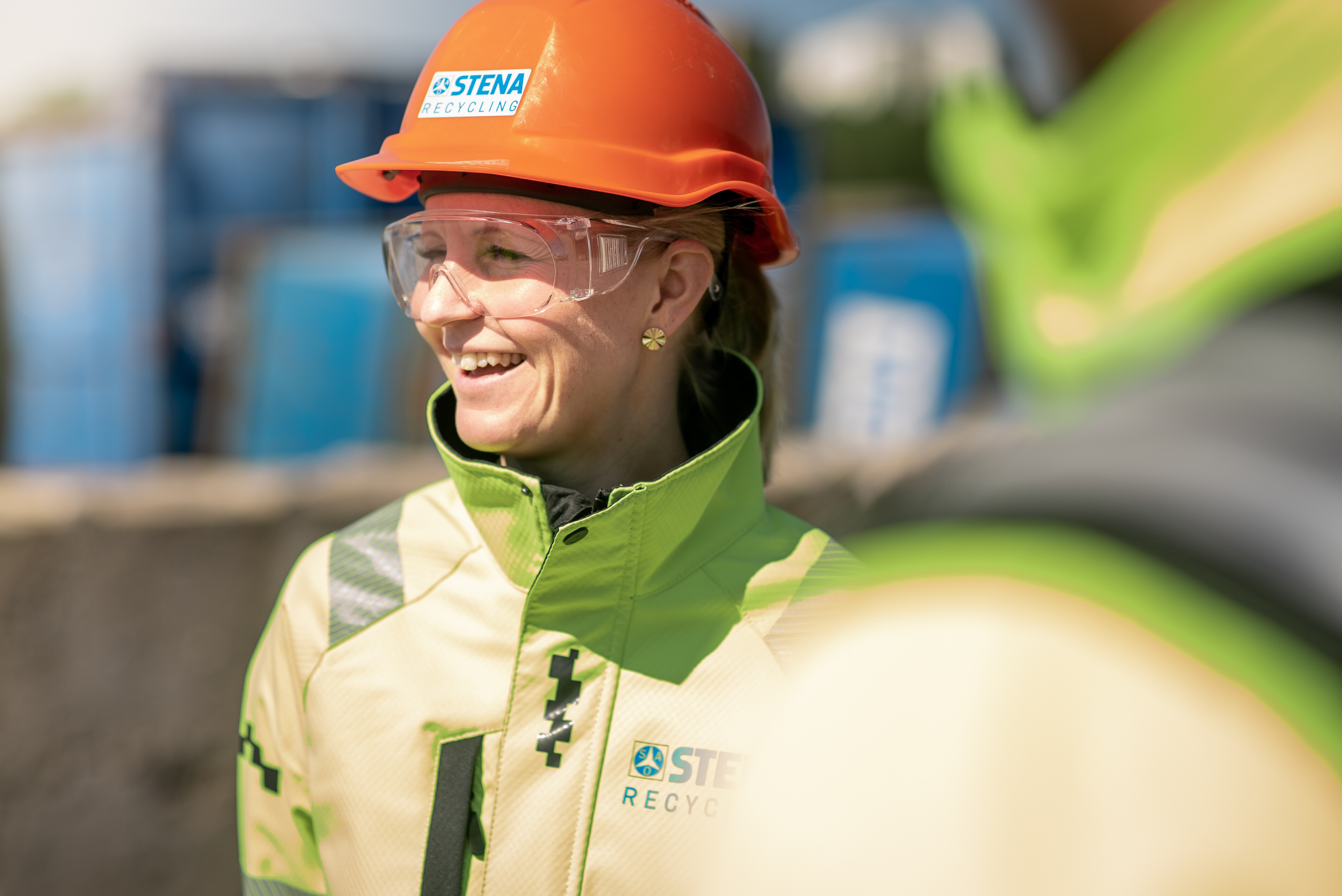 Person in Stena Recycling safety gear and orange helmet standing near blue industrial containers.
