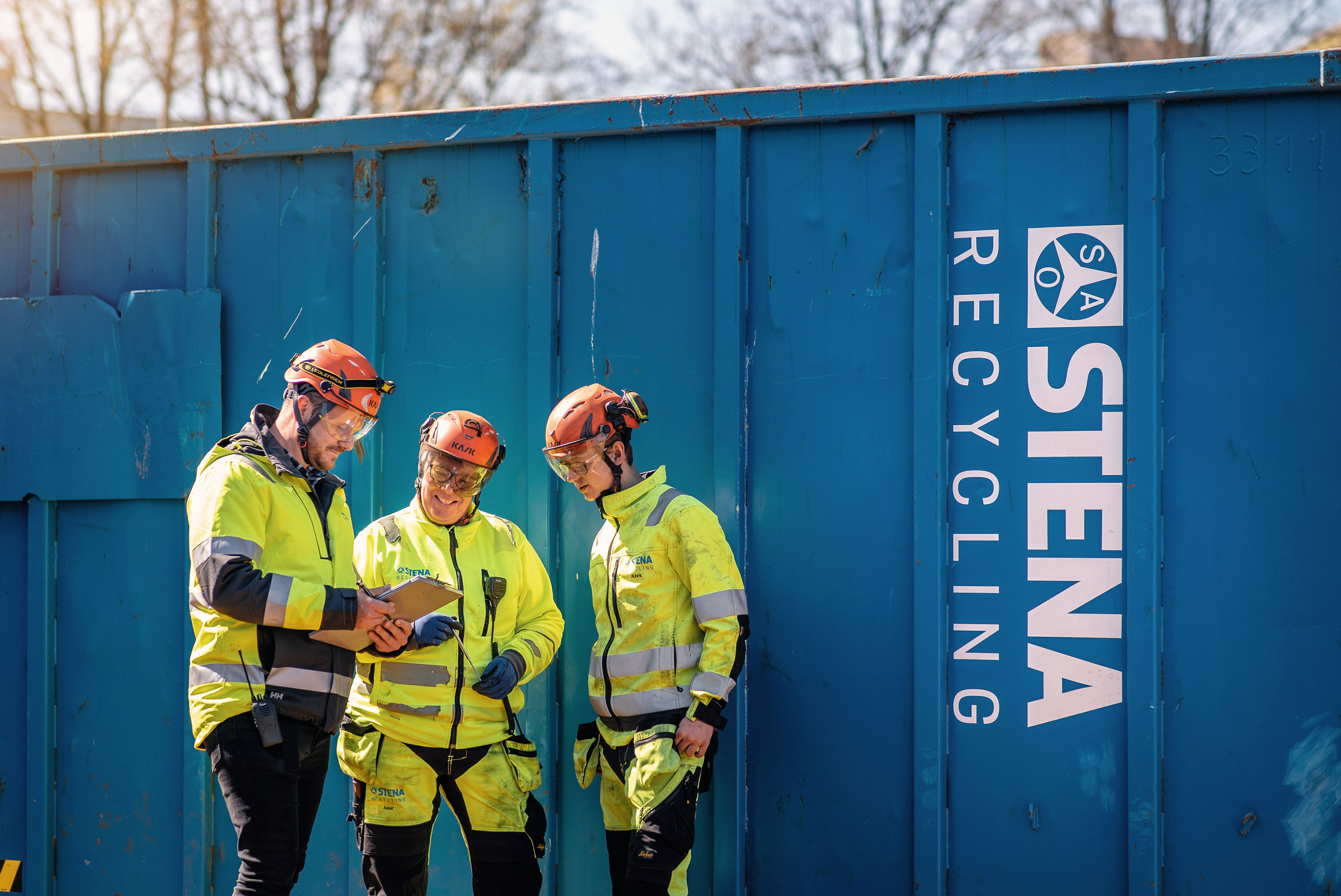 Three Stena Recycling employees in protective, hi-vis gear look at a clipboard beside a scrap collection container.