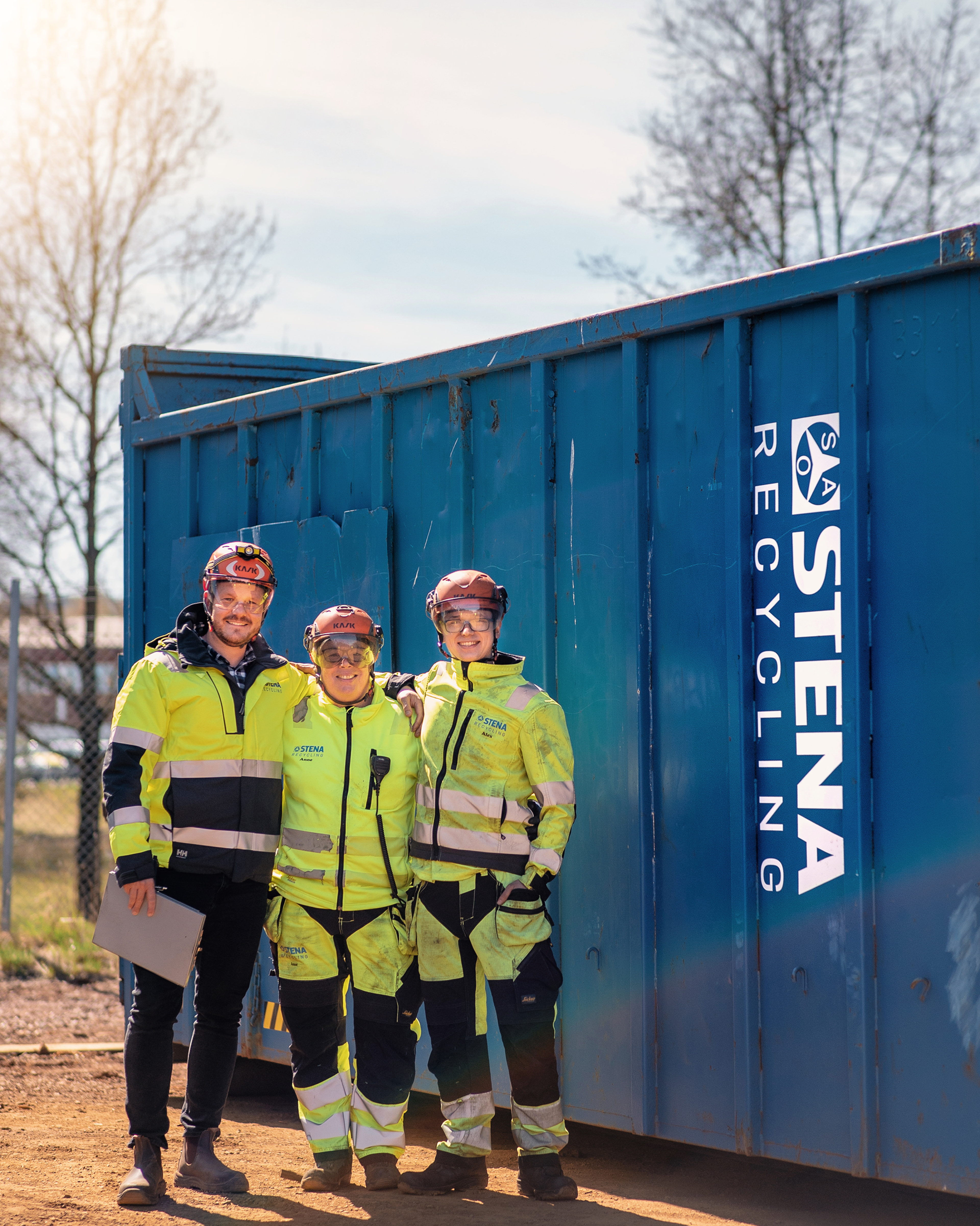 Tre experter på Stena Recycling klädda i reflekterande arbetskläder, hjälmar och skyddsglasögon, står framför en blå Stena Recycling stålcontainer med armarna om varandra och ler mot fotografen