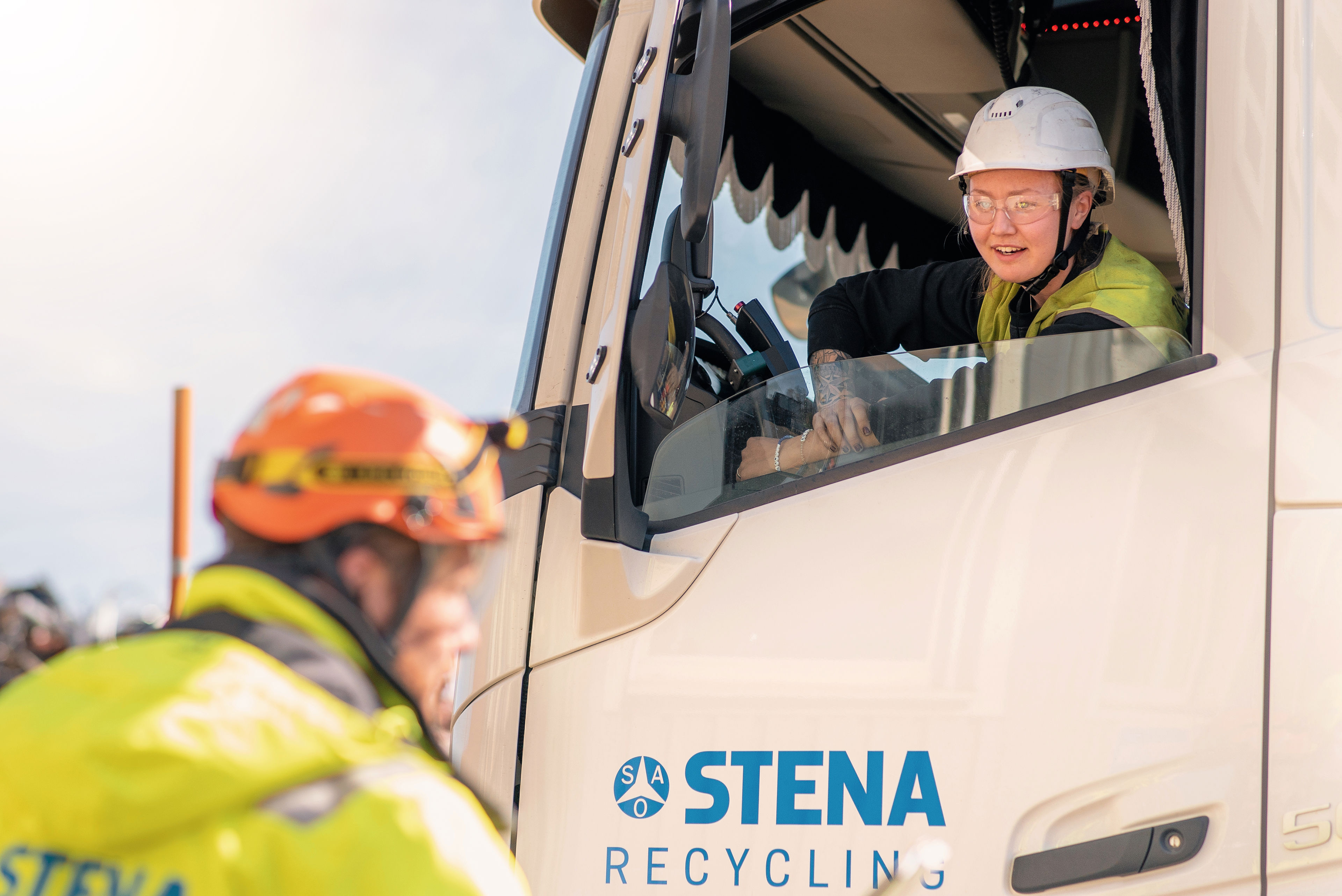 A female Stena Recycling employee driving a waste collection truck talks with a male colleague standing nearby.