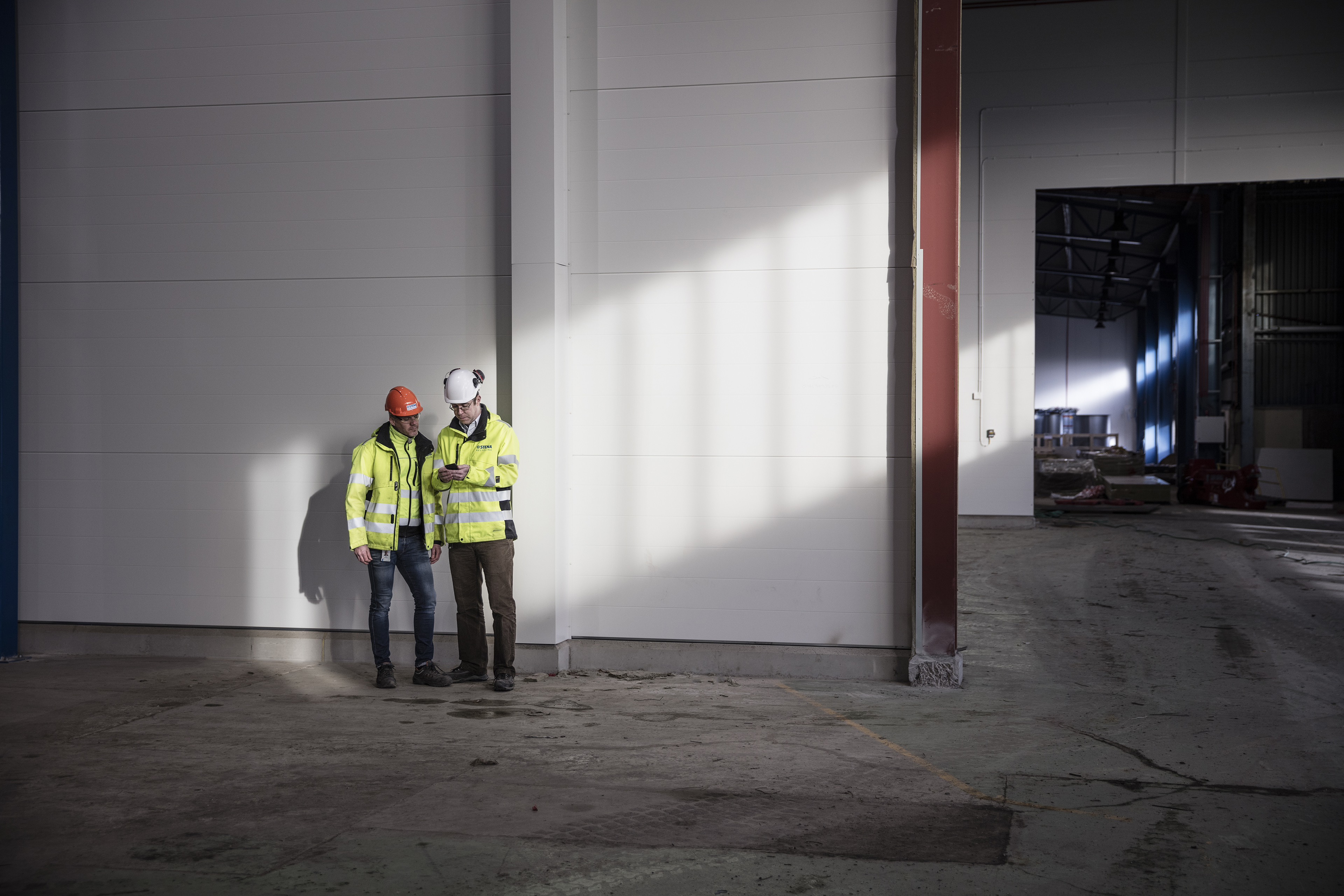Two male Stena Recycling employees in protective gear look at a mobile phone together in a recycling facility