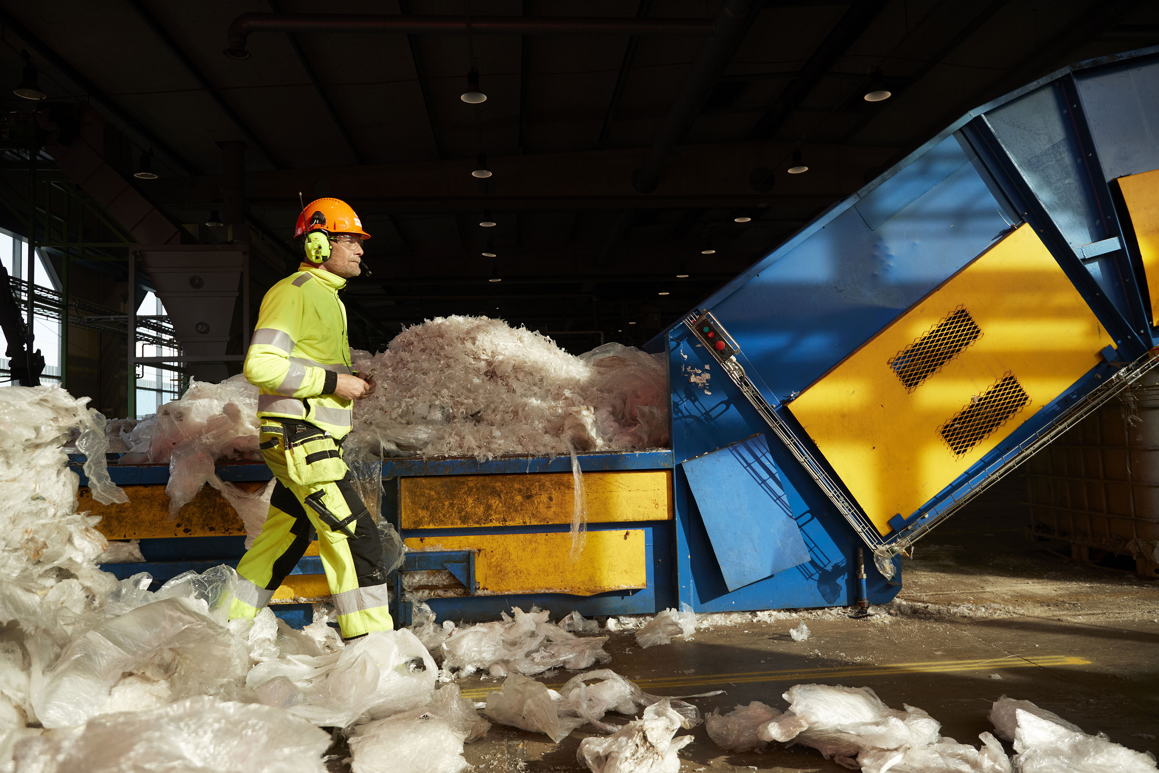 Återvinningsexpert som är klädd i reflekterande arbetskläder, bär hjälm, hörselskydd och skyddsglasögon, passerar en maskin som återvinner plast.