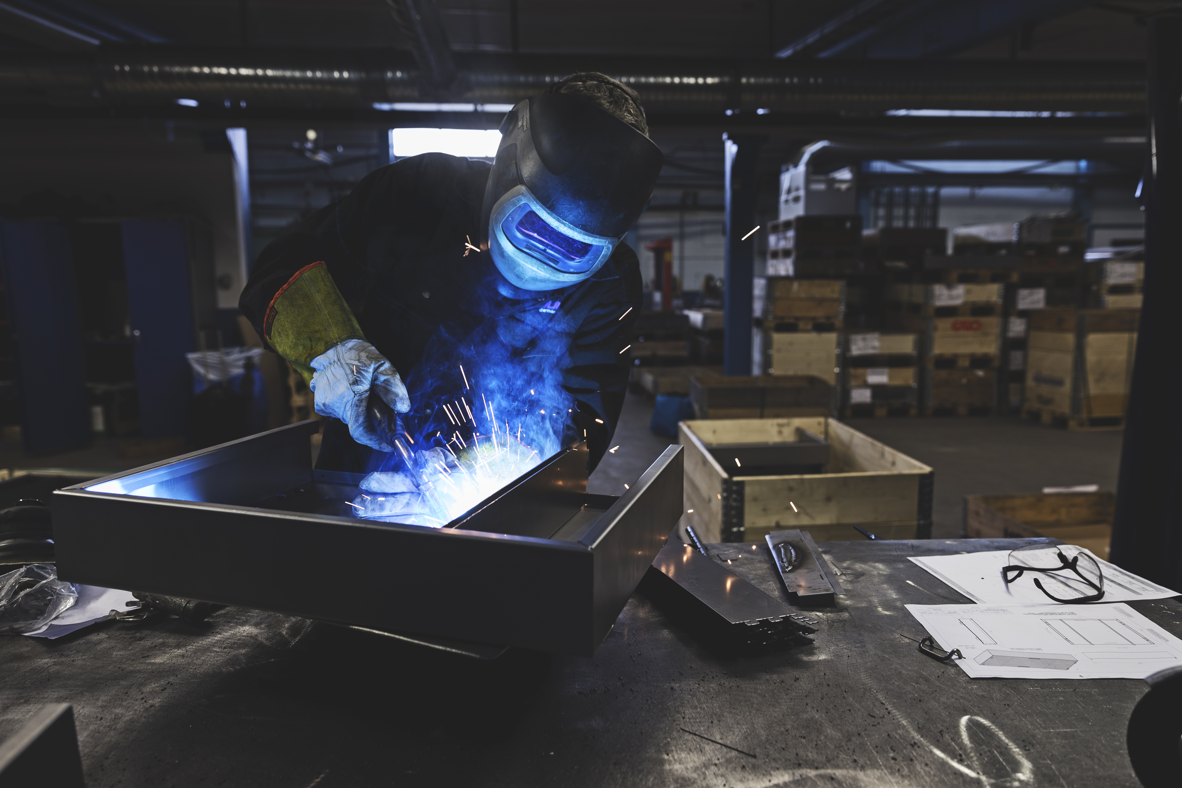 Ein Arbeiter in Schutzausrüstung schweißt ein Metallstück in einer Werksumgebung.