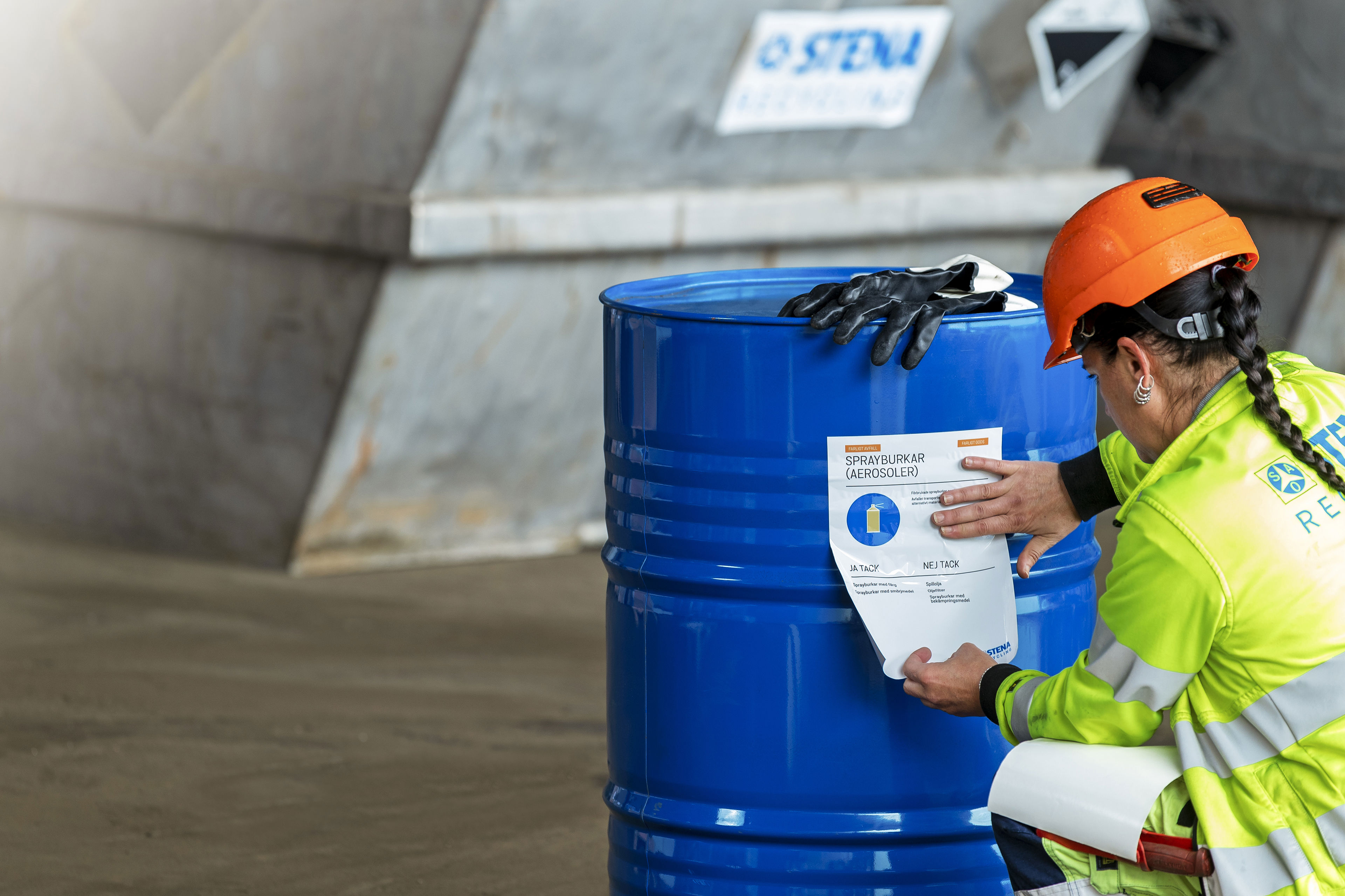 A Stena Recycling expert dressed in hardhat and reflective work jacket checking the safety label on a blue steel barrel containing hazardous waste for recycling.